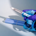 Hand holding Ozempic 1 mg semaglutide GLP-1 pens with a blue measuring tape on a blue gradient background.