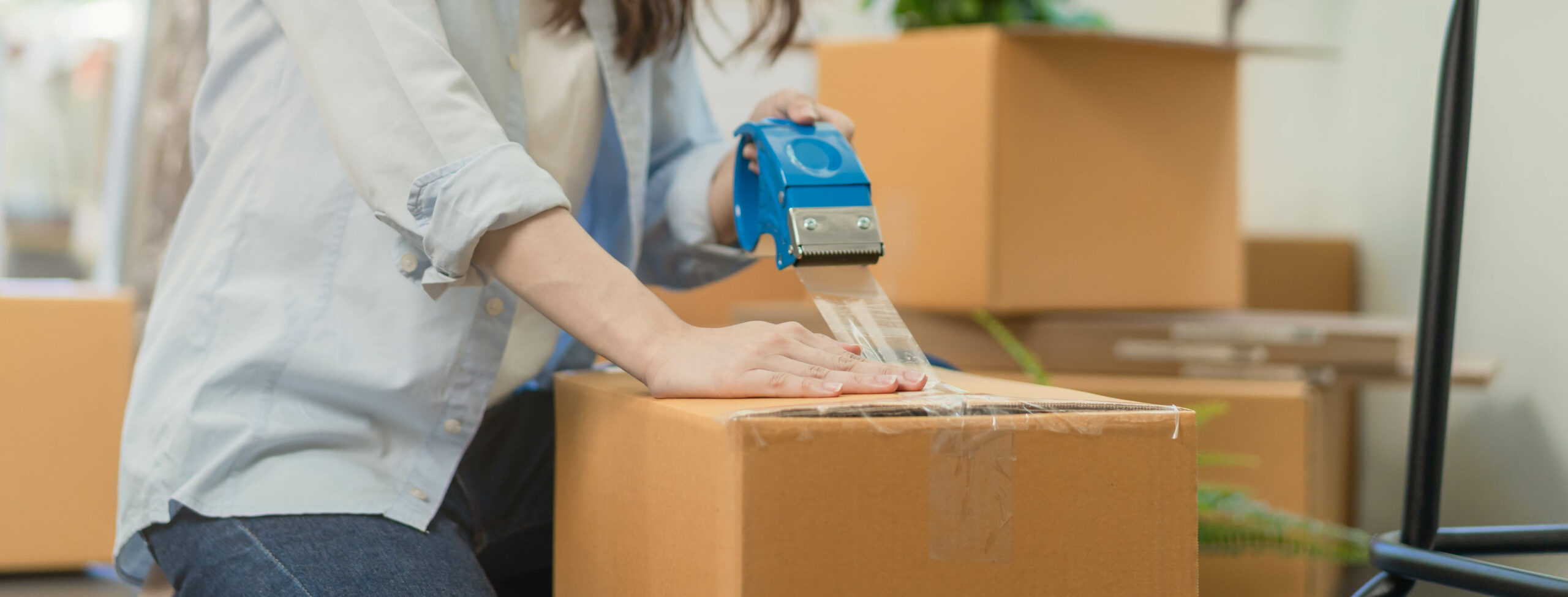 Hand of woman holding packing box by tape machine, sealing cardboard indoors.