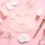 Facial sheet mask with different cosmetic products and gypsophila flowers on pink background