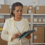 Woman adjusts glasses while holding a tablet amid stacked cardboard boxes on shelves in a storage building.
