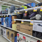 A selection of household appliances including vacuum cleaners steam irons and small home electronics are displayed with price tags inside a grocery store.