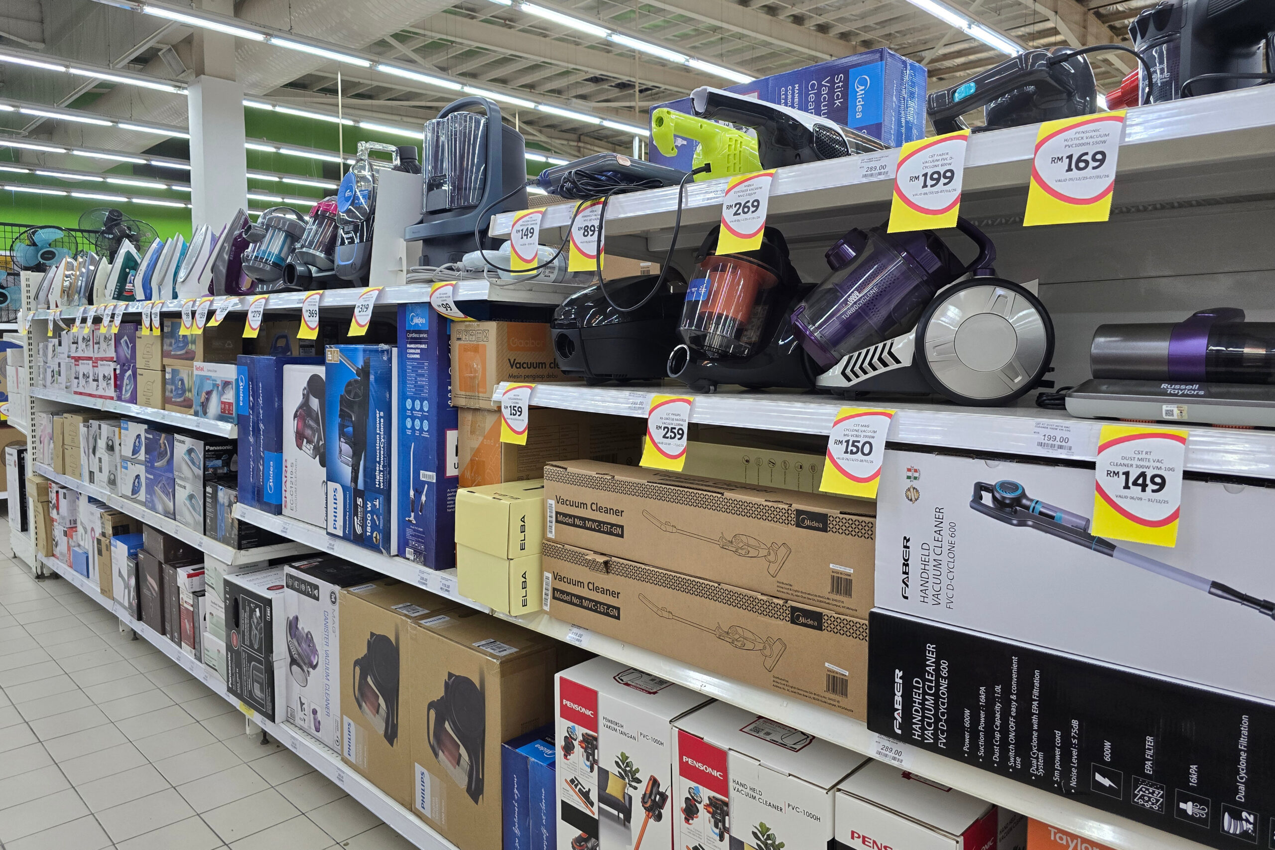 A selection of household appliances including vacuum cleaners steam irons and small home electronics are displayed with price tags inside a grocery store.