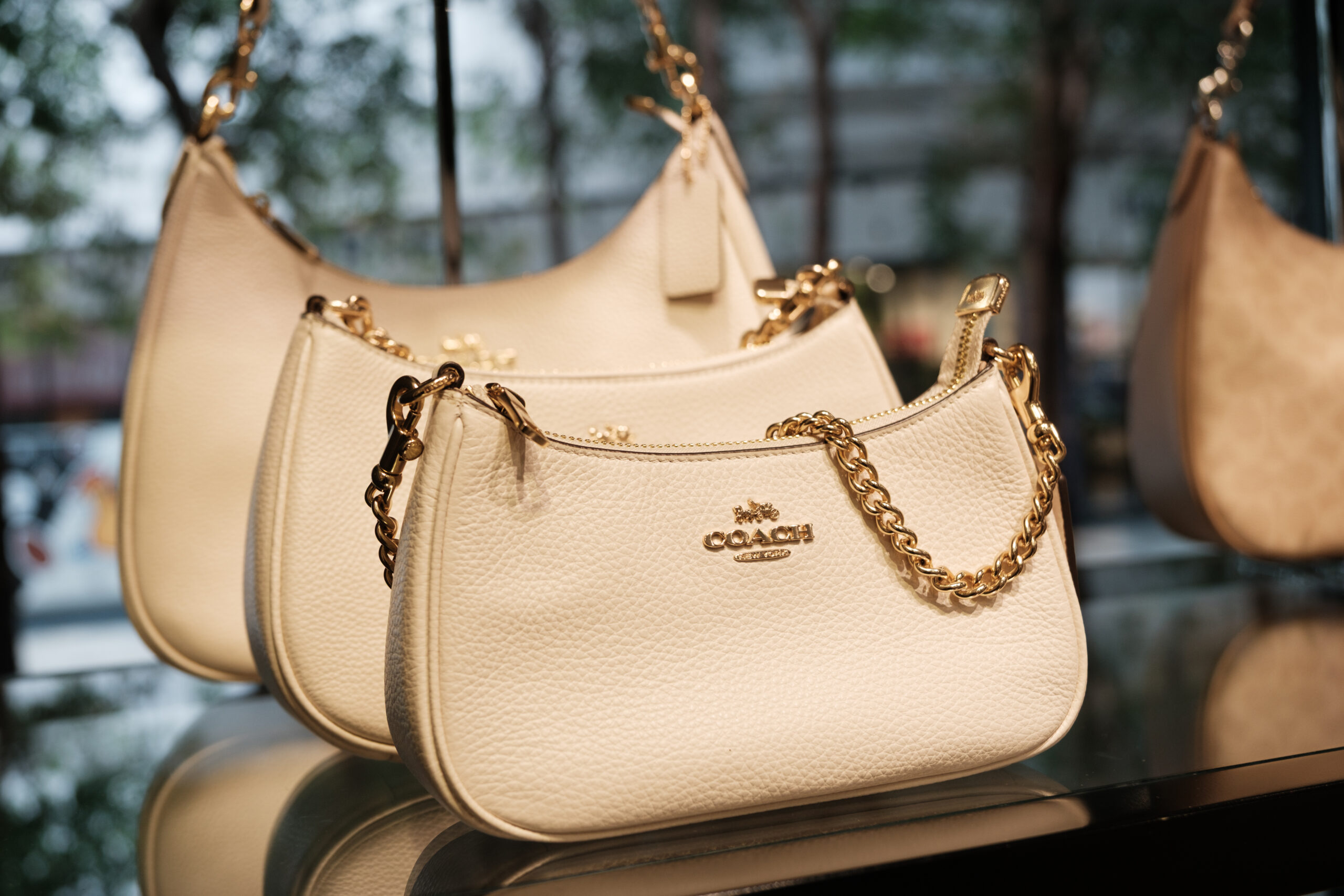 Coach branded leather handbags in white are arranged on display, featuring gold chain details and a shiny logo against a blurred outdoor backdrop.