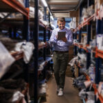 Full length of supervisor walking trough storage of export firm, checking on goods and using tablet.
