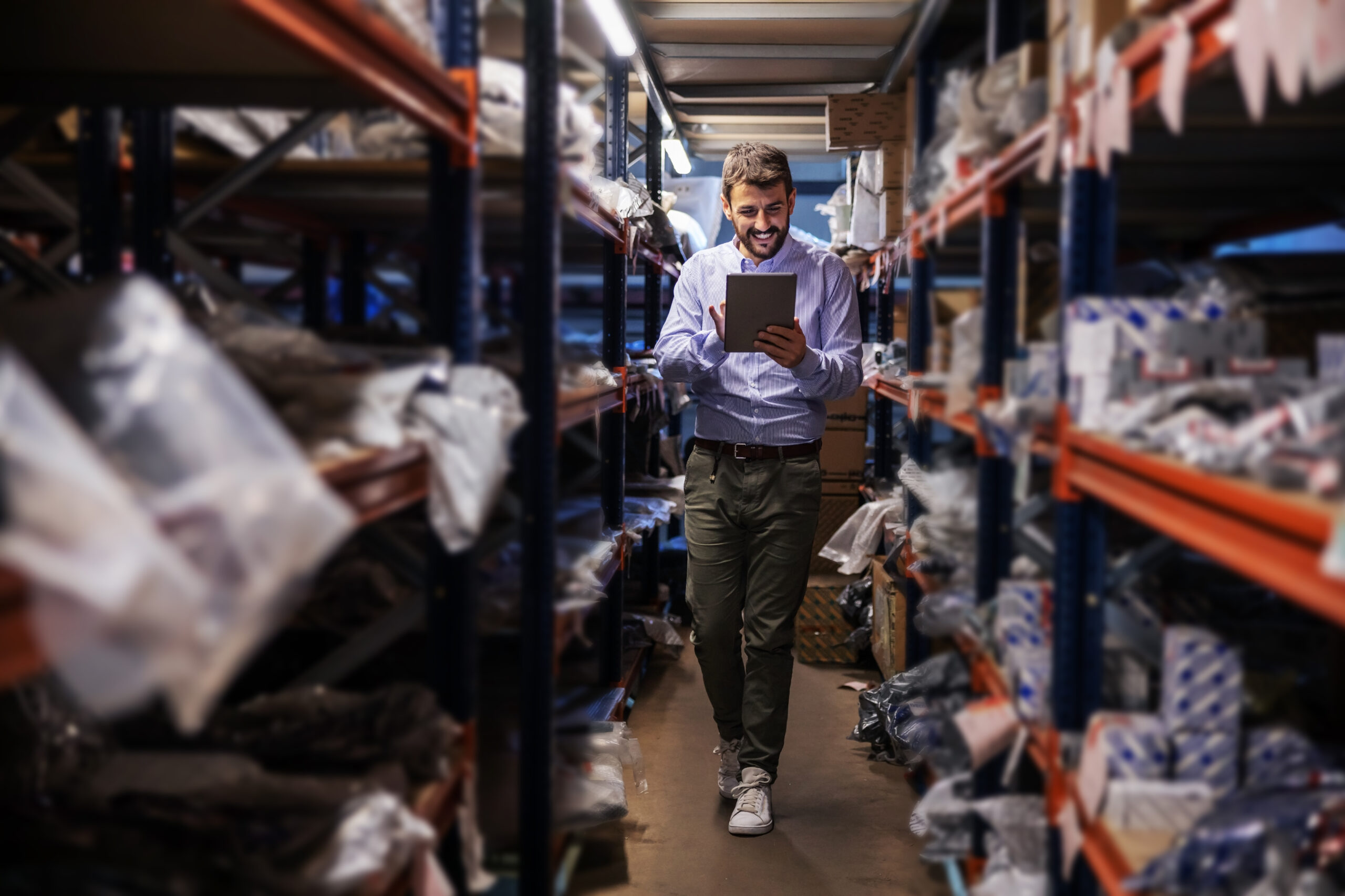 Full length of supervisor walking trough storage of export firm, checking on goods and using tablet.