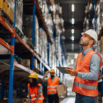 Warehouse manager holding digital tablet, looking up at shelves while checking inventory with his team in the background