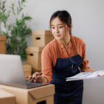 female business owner managing online store operations, checking orders and inventory on laptop while preparing packages for e-commerce shipping