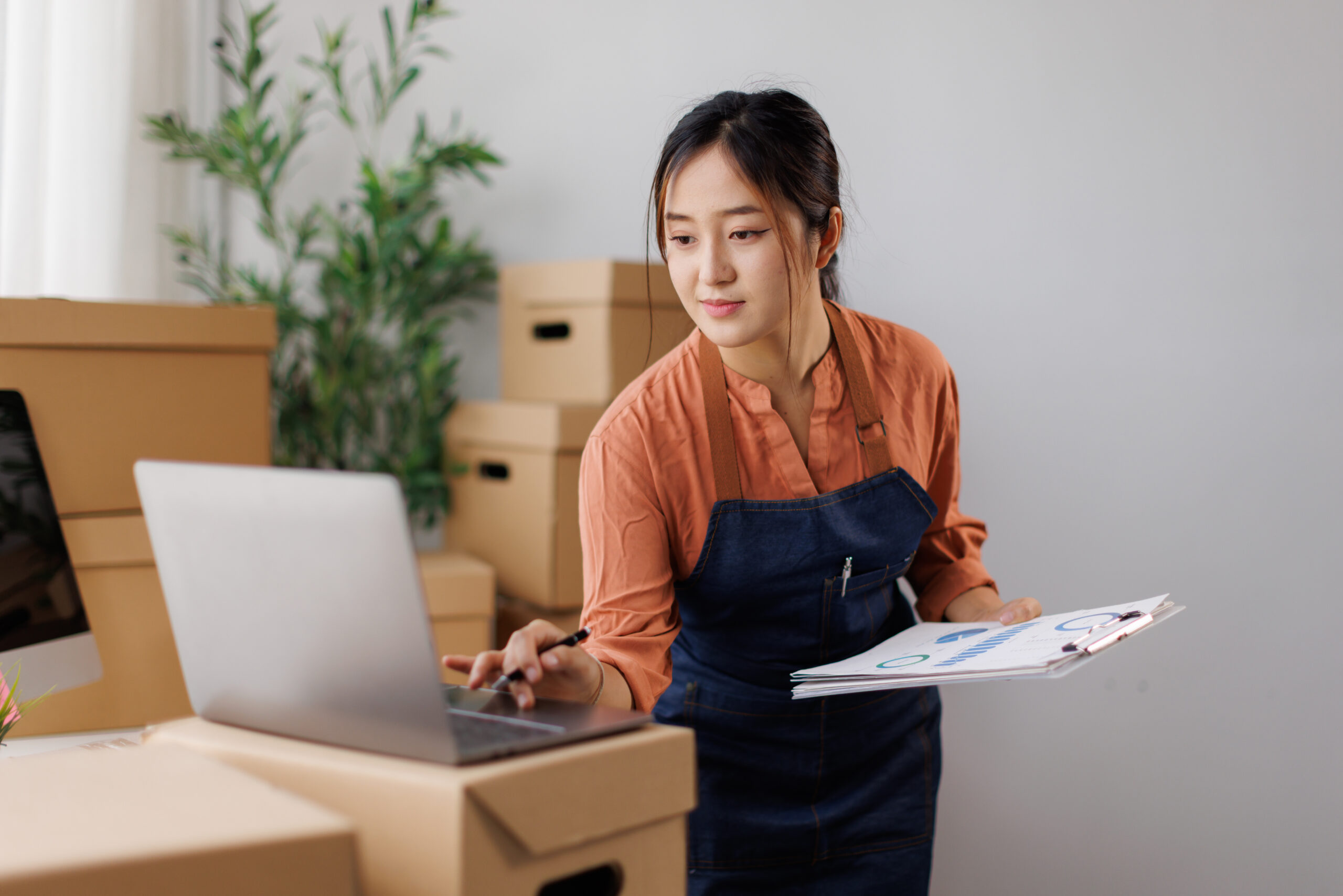 female business owner managing online store operations, checking orders and inventory on laptop while preparing packages for e-commerce shipping