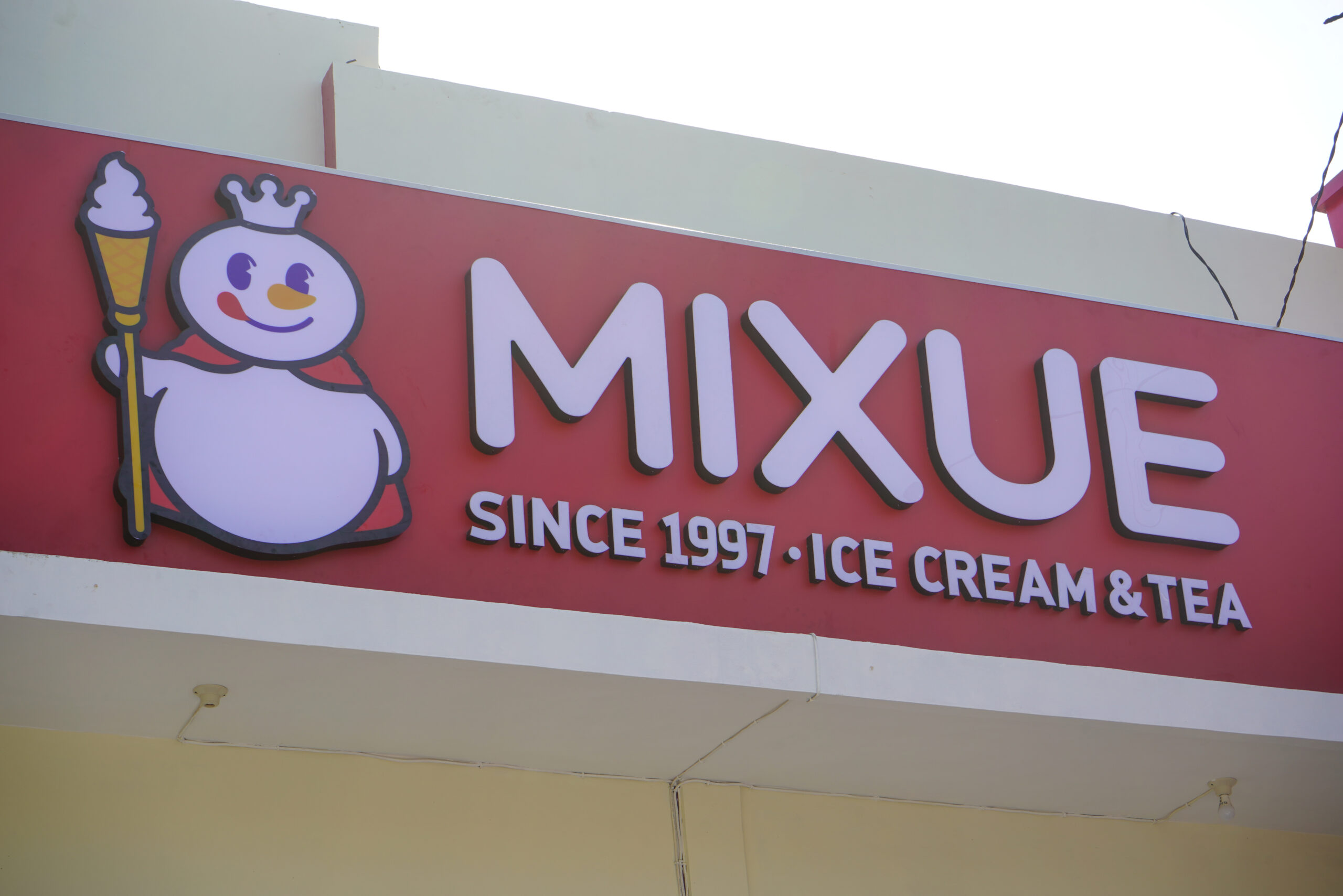 Mixue Ice Cream and Tea shop storefront sign featuring the iconic snowman mascot and brand name in red and white background, captured outdoors during the day in a modern urban commercial building.