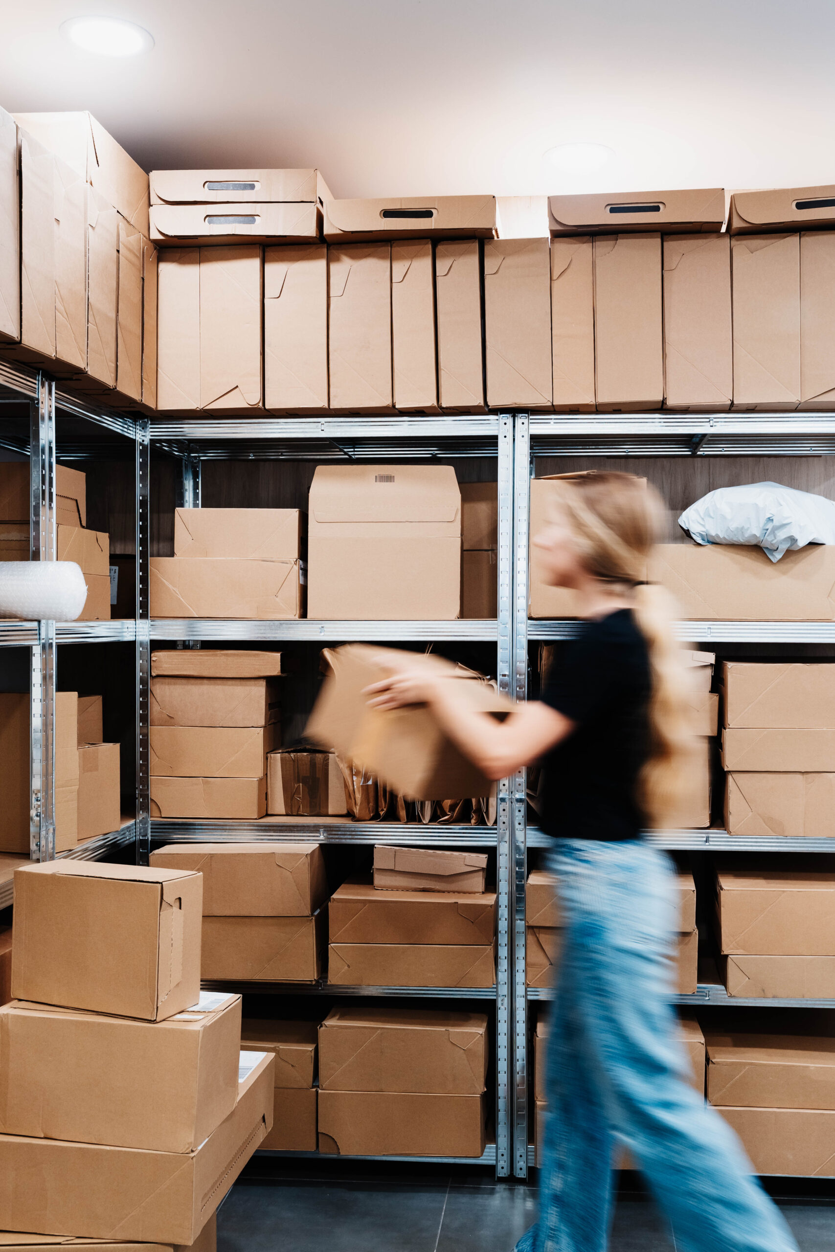 Blurred woman in motion pulls a cardboard box from metal shelving in a small storage room packed with parcels.
